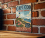A vintage metal sign for Cape Cod, Massachusetts, featuring a lighthouse and a beach scene.