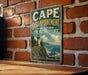 A vintage-style metal sign with the text "CAPE DISAPPOINTMENT STATE PARK WASHINGTON" and an illustration of a lighthouse on a cliff overlooking the ocean.