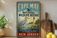 A vintage-style metal sign for Cape May National Wildlife Refuge, featuring a blue heron and a lighthouse, is displayed on a shelf next to a wooden cutting board and some lemons.