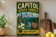 A vintage-style metal sign for Capitol State Forest in Washington, featuring a mountain landscape with pine trees and a river, along with a sign that says "Explore Discover Protect Washington".