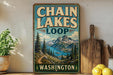 A vintage-style metal sign with the text "CHAIN LAKES LOOP WASHINGTON" displayed on a wooden shelf, with a scenic mountain landscape in the background.