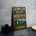 A vintage metal sign for the Champlain Valley National Heritage Partnership, featuring a scenic landscape with a lighthouse and boats, stands on a gray countertop next to a stack of black camera lenses.