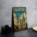 A vintage metal sign displaying the name "CHARTRES Cathedral" and the word "FRANCE" is placed on a gray countertop next to a stack of black camera lenses.