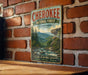 A vintage-style metal sign for the Cherokee National Forest in Tennessee, featuring a scenic mountain landscape with pine trees and a river.