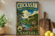 A vintage-style sign for Chickasaw State Forest in Tennessee, featuring a scenic landscape with pine trees, a river, and mountains.