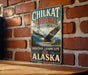 A vintage metal sign with a bald eagle and the words "CHILKAT BALD EAGLE PRESERVE" is displayed on a brick wall.