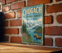 A vintage-style metal sign with the text "CHUGACH NATIONAL FOREST ALASKA" and an illustration of a moose in a forest setting, mounted on a brick wall.