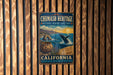 A metal sign with a colorful illustration of a whale, a pot, and a coastal landscape, mounted on a wooden wall.