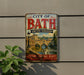A vintage metal sign on a wall, depicting a cityscape with a bridge and water, advertising the "City of Bath United Kingdom" and highlighting its "History, Heritage, Beauty".