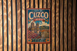 A vintage-style metal sign with the text "CITY OF Cuzco PERU" and an illustration of a historic Inca city, mounted on a wooden wall.