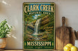 A vintage-style metal sign for Clark Creek Natural Area in Mississippi, featuring a waterfall and lush greenery.