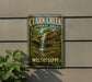 A vintage-style metal sign for Clark Creek Natural Area in Mississippi, featuring a waterfall and lush greenery.