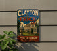 A vintage metal sign with the text "CLAYTON THE FRICK PITT'SBURGH PENNSYLVANIA" displayed on a wall, with a building and landscaping in the background.