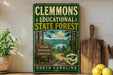 A vintage-style metal sign for Clemmons Educational State Forest in North Carolina, featuring a scenic landscape with a lake and pine trees.