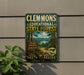 A metal sign for Clemmons Educational State Forest in North Carolina, featuring a scenic image of a forest trail and a lake.