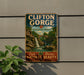 A vintage-style metal sign for Clifton Gorge, Ohio, featuring a waterfall and lush greenery, mounted on a gray wall.