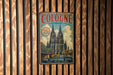 A vintage metal sign with the text "COLOGNE GERMANY" and an illustration of the Cologne Cathedral, hanging on a wooden wall.