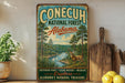 a vintage-style sign for Conecuh National Forest, Alabama, with a scenic illustration of a forest and a river. The sign is displayed on a shelf next to a wooden cutting board and a potted plant.