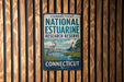 A vintage metal sign for the Connecticut National Estuarine Research Reserve, featuring a seagull and a lighthouse, is displayed on a wooden wall.
