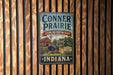 A vintage-style metal sign for the Conner Prairie Living History Museum in Indiana, featuring an illustration of a horse-drawn carriage and a barn.