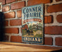 A vintage-style metal sign for the Conner Prairie Living History Museum in Indiana, featuring an illustration of a horse-drawn carriage and a red barn.