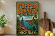 A vintage-style sign for Copper Country State Forest in Michigan, featuring a scenic landscape with pine trees, a river, and mountains.