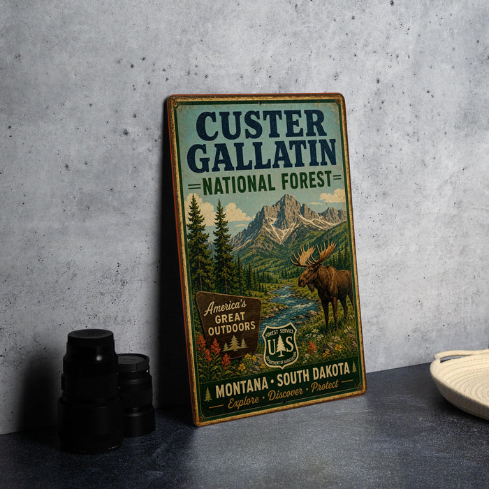 A vintage-style sign for Custer Gallatin National Forest, featuring a moose and mountains in the background, placed on a gray countertop next to some black camera lenses.