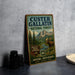 A vintage-style sign for Custer Gallatin National Forest, featuring a moose and mountains in the background, placed on a gray countertop next to some black camera lenses.