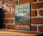 A vintage-style sign for "Yukon" is displayed on a brick wall, featuring a scenic mountain landscape with pine trees and a river.