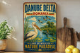 A vintage-style sign for Danube Delta, Romania, featuring a white stork and a boat on a river surrounded by lush greenery.