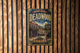 A vintage-style metal sign for Deadwood Saloon, featuring an illustration of a saloon building and a horse-drawn carriage, mounted on a wooden wall.