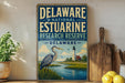 A vintage-style sign for the Delaware National Estuarine Research Reserve, featuring an illustration of a heron and a lighthouse, is displayed on a shelf next to a wooden cutting board and some lemons.