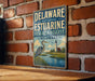 A vintage metal sign for the Delaware National Estuarine Research Reserve, featuring a blue heron and a lighthouse, is displayed on a brick wall.