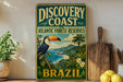 A vintage-style metal sign advertising the Discovery Coast Atlantic Forest Reserve in Brazil, featuring a colorful illustration of a toucan and tropical foliage.