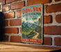 A vintage-style metal sign with a scenic mountain landscape and a winding road, mounted on a brick wall.