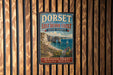 A vintage-style metal sign with text and a scenic image of the Dorset and East Devon Coast in the United Kingdom, hanging on a wooden wall.