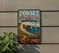 A vintage-style metal sign advertising the Dorset and East Devon Coast in the United Kingdom, featuring a scenic coastal landscape with cliffs, a beach, and a sailboat.