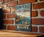 A vintage-style metal sign with the text "Down East Coastal Heritage Landscape" and "Wild Coast, Rich Heritage, Timeless Beauty" is displayed on a brick wall.
