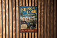 A vintage advertisement poster for Edison and Ford, featuring two men in suits and a classic car, is displayed on a wooden wall.
