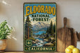 A vintage-style metal sign for Eldorado National Forest in California, featuring a scenic landscape with mountains, pine trees, and a river.