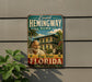 A vintage-style sign for Ernest Hemingway's home in Key West, Florida, featuring a painting of a man and a cat in a tropical setting.