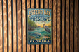 A vintage-style metal sign for Estero Bay Aquatic Preserve in Florida, featuring an illustration of various marine life and a bird.