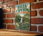 A vintage metal sign for Falls Canyon, Indiana, is displayed on a brick wall.