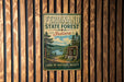 A vintage-style metal sign for the Ferdined State Forest in Indiana, featuring a scenic landscape with pine trees, a lake, and mountains.