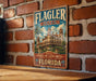A vintage-style sign for the Flagler Museum, featuring an illustration of a historic hotel with palm trees and a cloudy sky.