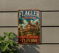A vintage-style metal sign for the Flagler Museum in Florida, featuring an illustration of a historic hotel with palm trees and a cloudy sky.