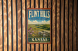 A vintage-style sign for Flint Hills, Kansas, featuring a scenic landscape with rolling hills, flowers, and a blue sky with clouds.