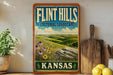 A vintage-style sign for Flint Hills, Kansas, featuring a scenic landscape with rolling hills, flowers, and a blue sky with clouds.