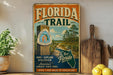 A vintage-style sign for the Florida Trail, featuring a scenic illustration of a trail surrounded by pine trees and a house, along with text promoting the trail as a hiking and exploring destination in Florida.