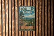 A vintage metal sign for the Foothills Trail in South Carolina, featuring a scenic mountain landscape with a trail and a pine tree.